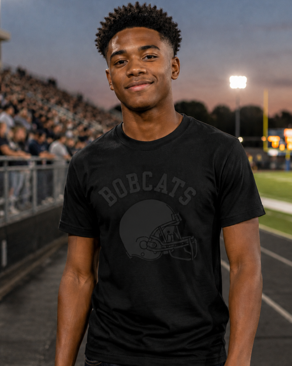 A young man standing on a school football field at dusk, wearing a black graphic t-shirt with black school mascot and a black football helmet design, paired with dark blue jeans.