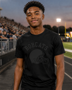 A young man standing on a school football field at dusk, wearing a black graphic t-shirt with black school mascot and a black football helmet design, paired with dark blue jeans.