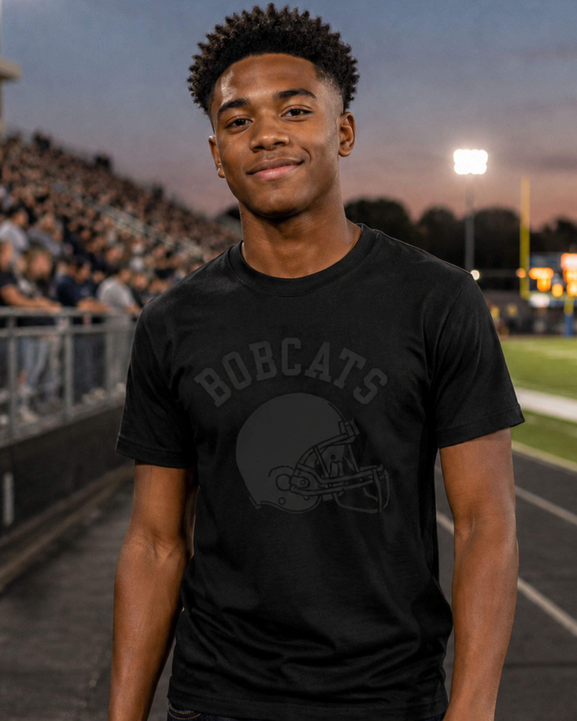 A young man standing on a school football field at dusk, wearing a black graphic t-shirt with black school mascot and a black football helmet design, paired with dark blue jeans.